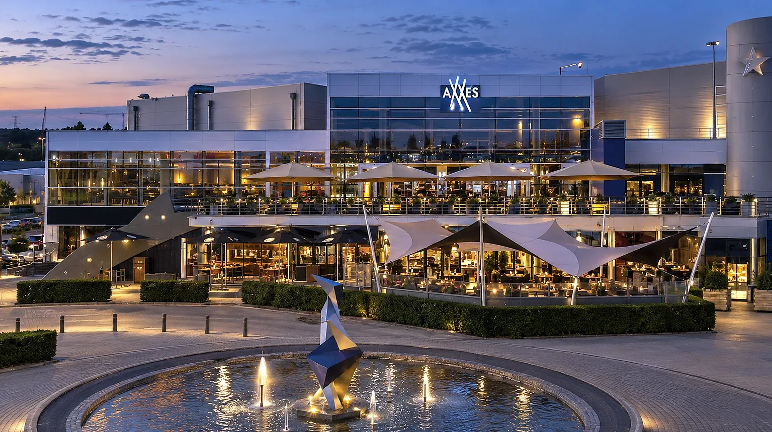 AXXES Café Restaurant bij avond: verlicht terras met parasols, de glazen gevel met het AXXES-logo en de fontein op het plein ervoor