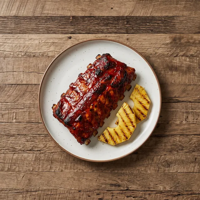 Spareribs van boven in een donkerrode marinade, met gegrilde ananas op een wit bord