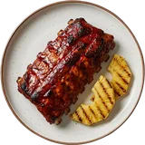 Spareribs van boven in een donkerrode marinade, met gegrilde ananas op een wit bord